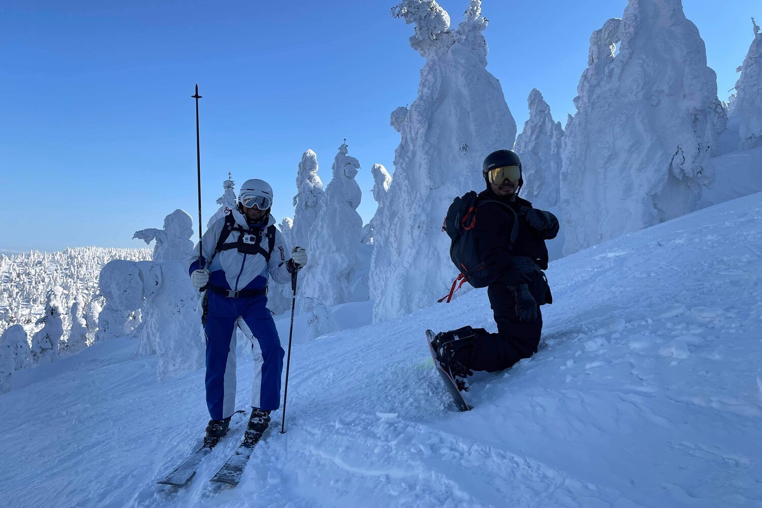 A snowboarder and skier dwarfed by Hakkoda's giant snow monsters, rime-frosted trees, on a bluebird day